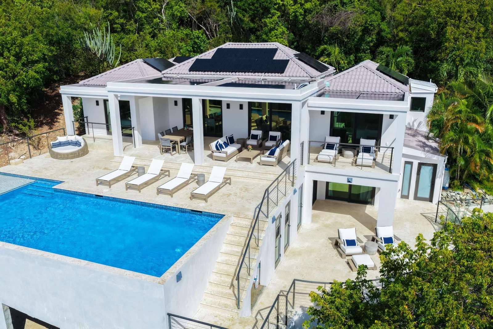 Multi-level pool deck with white loungers, teak outdoor sofas, dining table, and an infinity pool bordered by lush greenery. - property