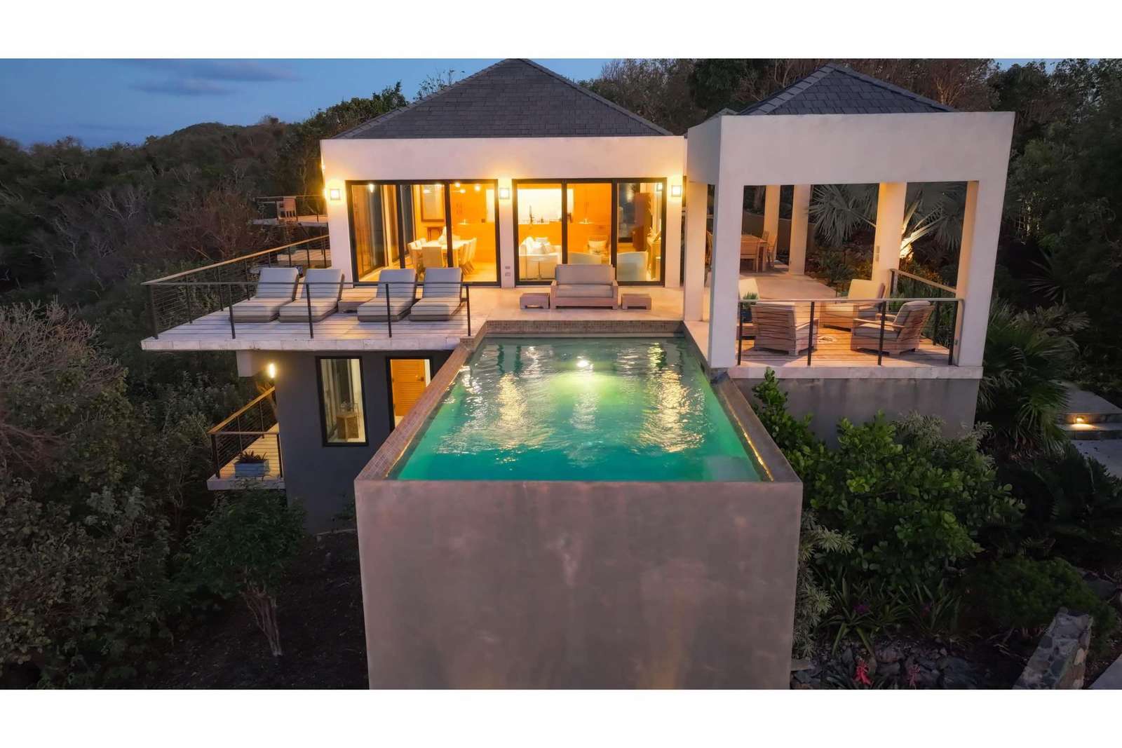 Infinity pool glows with underwater lights, bordered by a terrace with cushioned lounge chairs and covered seating. - property