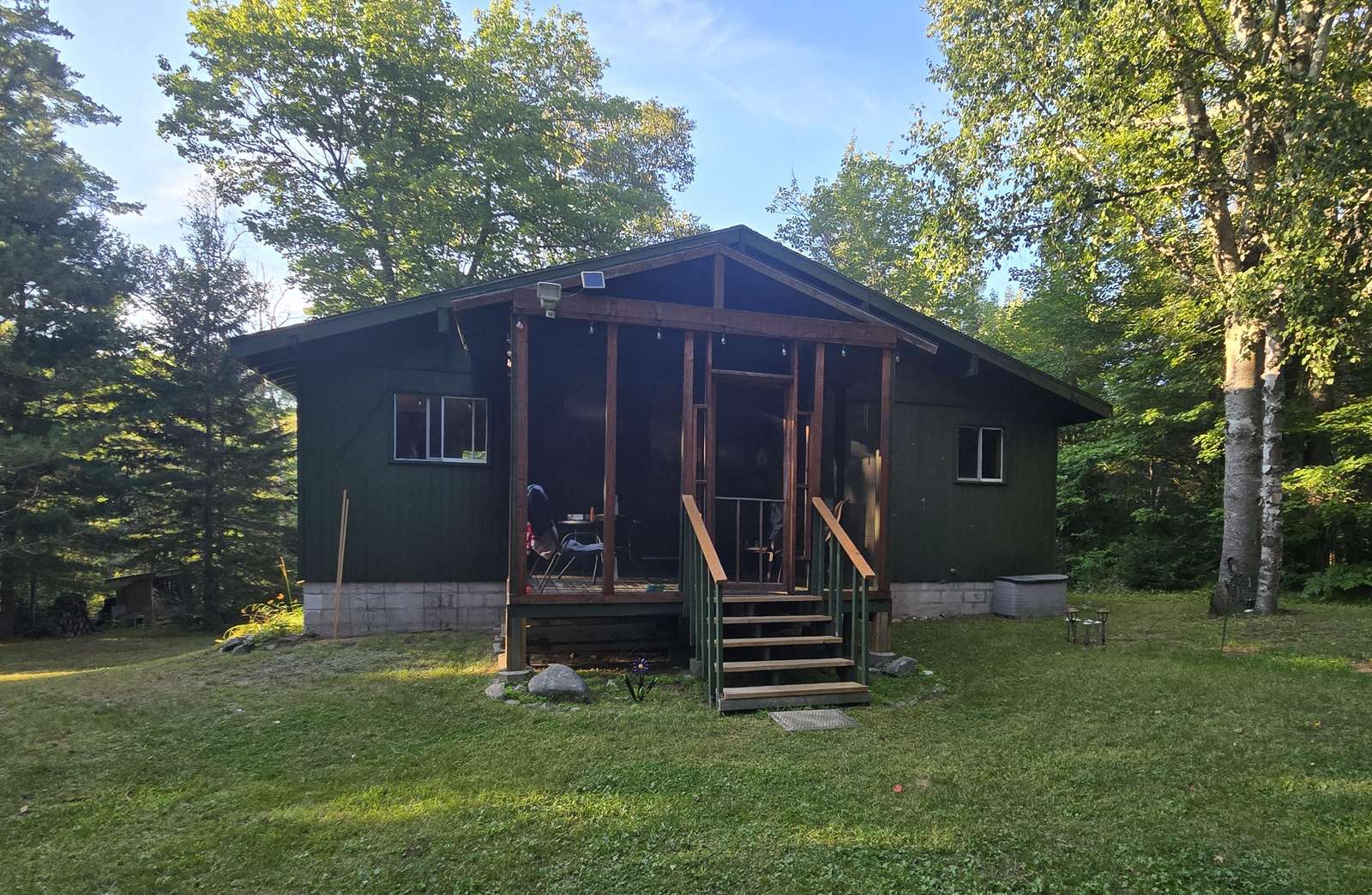 Exterior view of dark green cottage with screened porch entrance, wooden stairs, and surrounding mature trees. - property