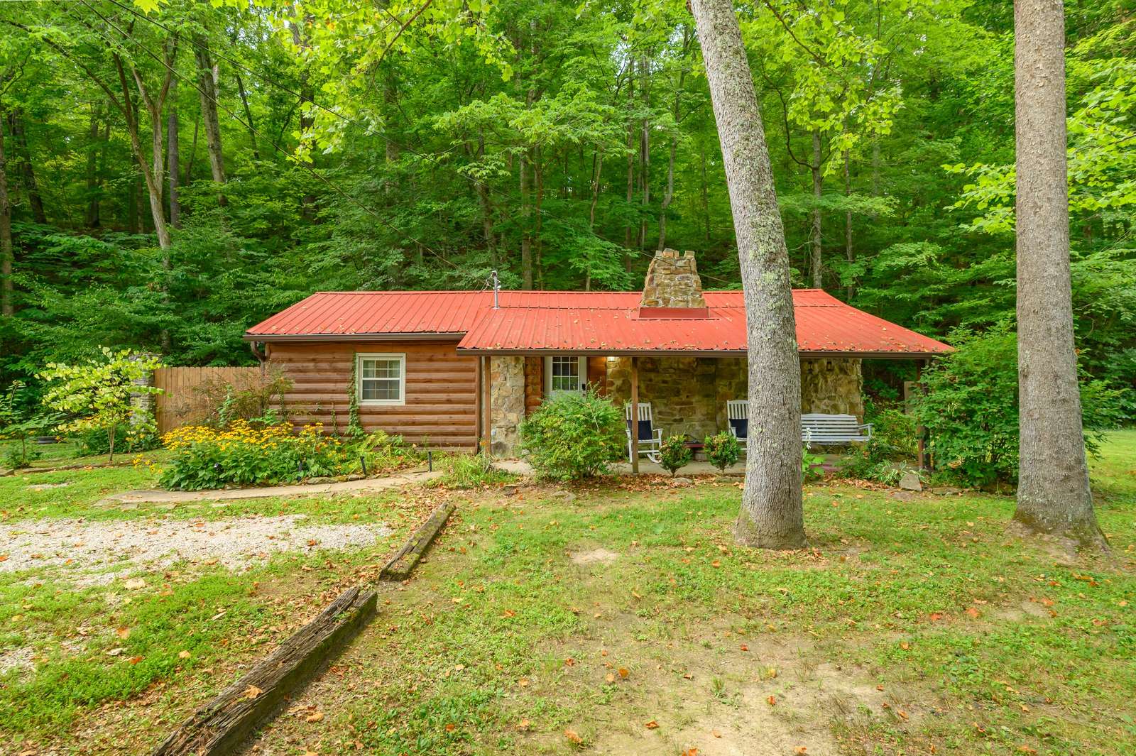 Front view of a rustic log cabin with a red metal roof, stone chimney, covered porch with rocking chairs, and lush green trees. - property