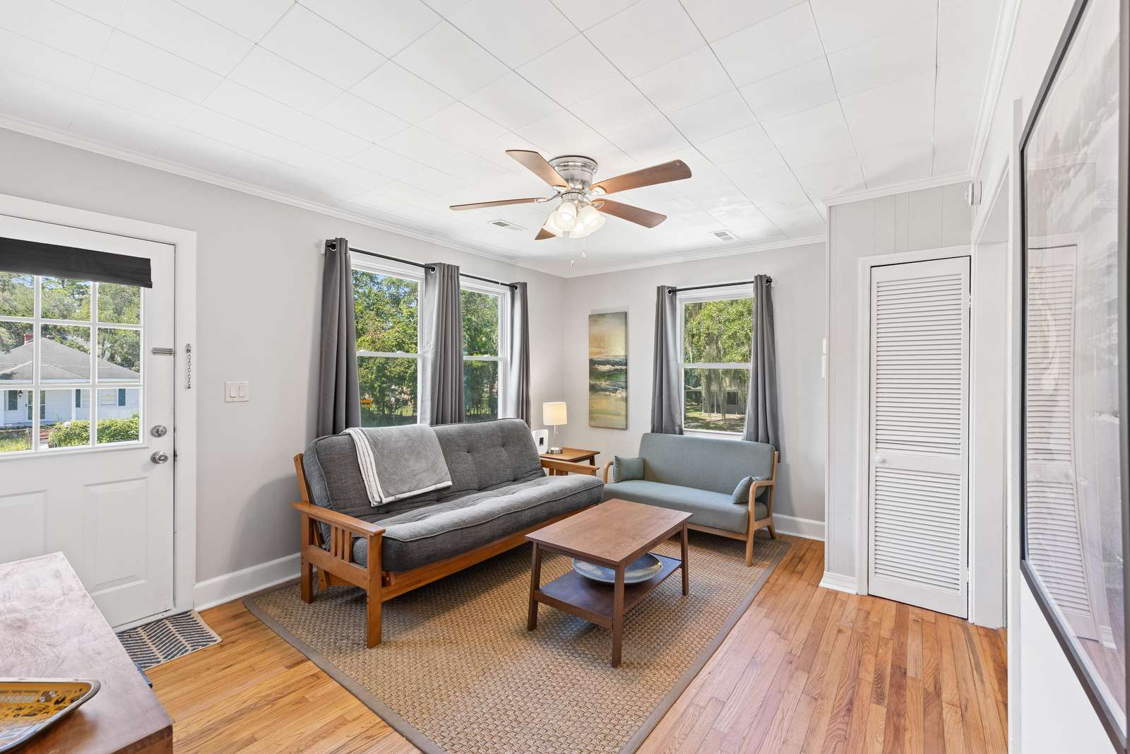 Natural light fills the living room through three windows framed by gray curtains, highlighting the hardwood floors and ceiling fan. - property