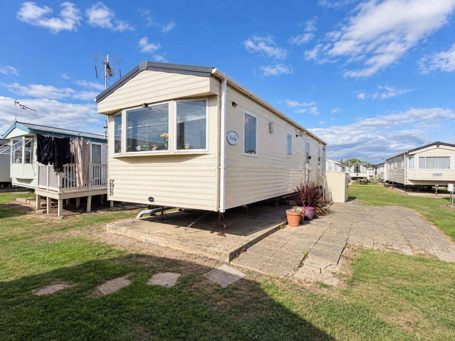 Exterior view of the cream mobile home with bay windows and potted plants on a stone patio under a blue sky. - property