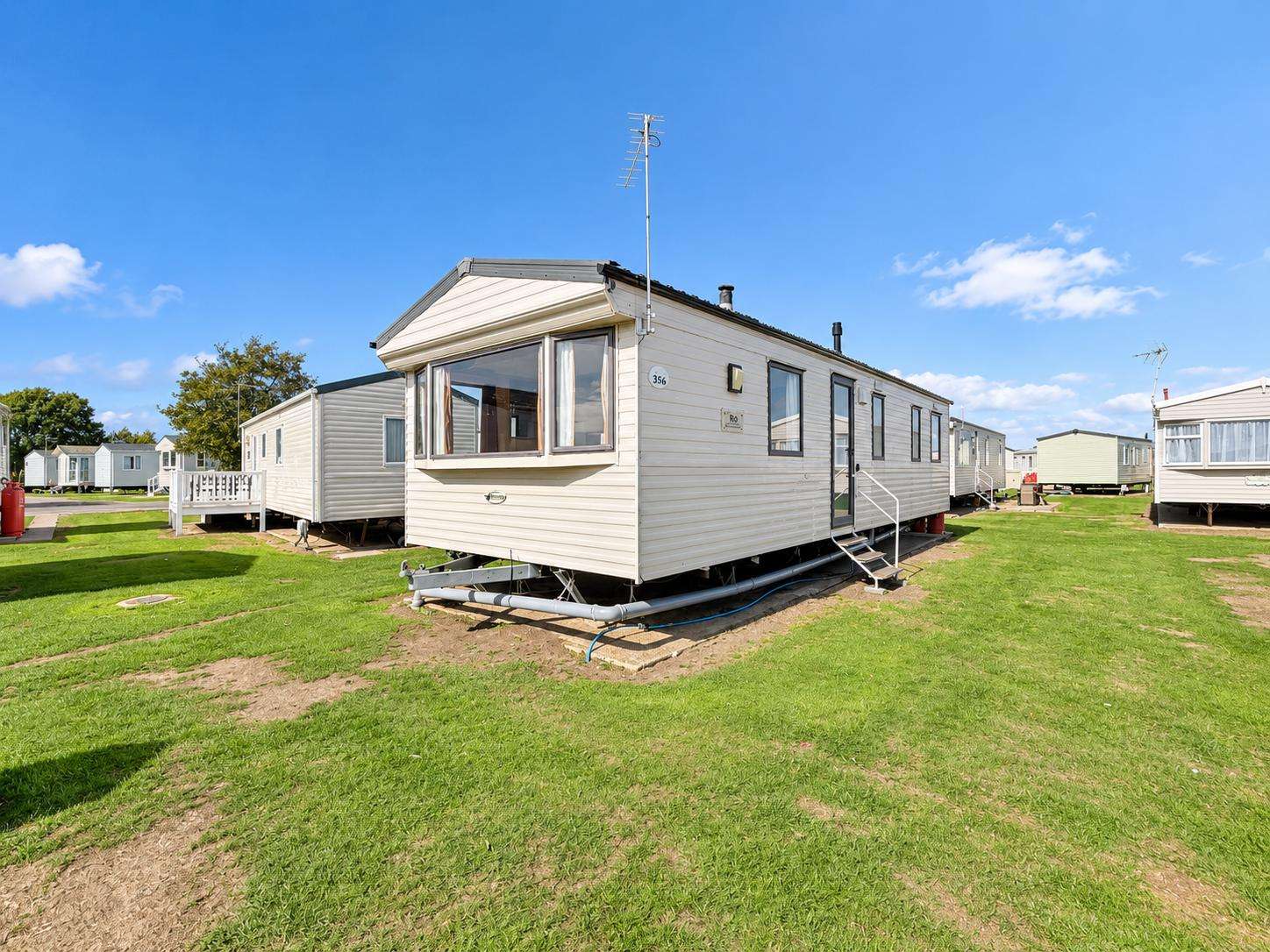 Exterior view of a cream-colored static caravan with large bay windows and metal steps on a green grassy plot. - property