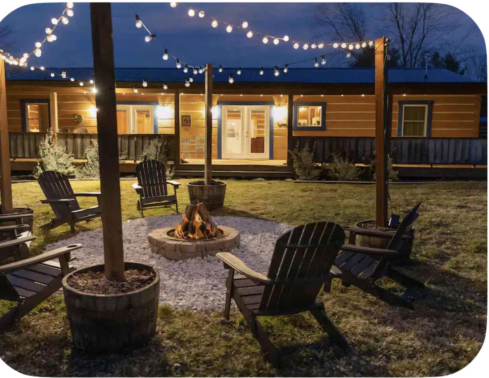 Outdoor fire pit encircled by dark wooden Adirondack chairs and string lights overhead at night. - property