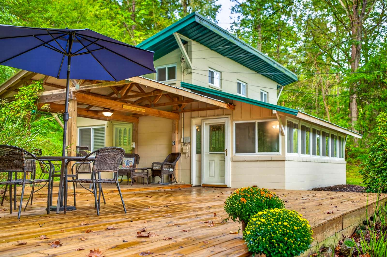 Spacious wooden deck with black wicker dining set, blue umbrella, and covered seating area against beige bungalow. - property