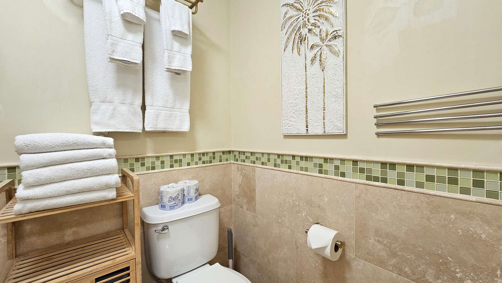 Bathroom corner with beige stone tiles, green mosaic border, white towels, and palm tree wall art. - property