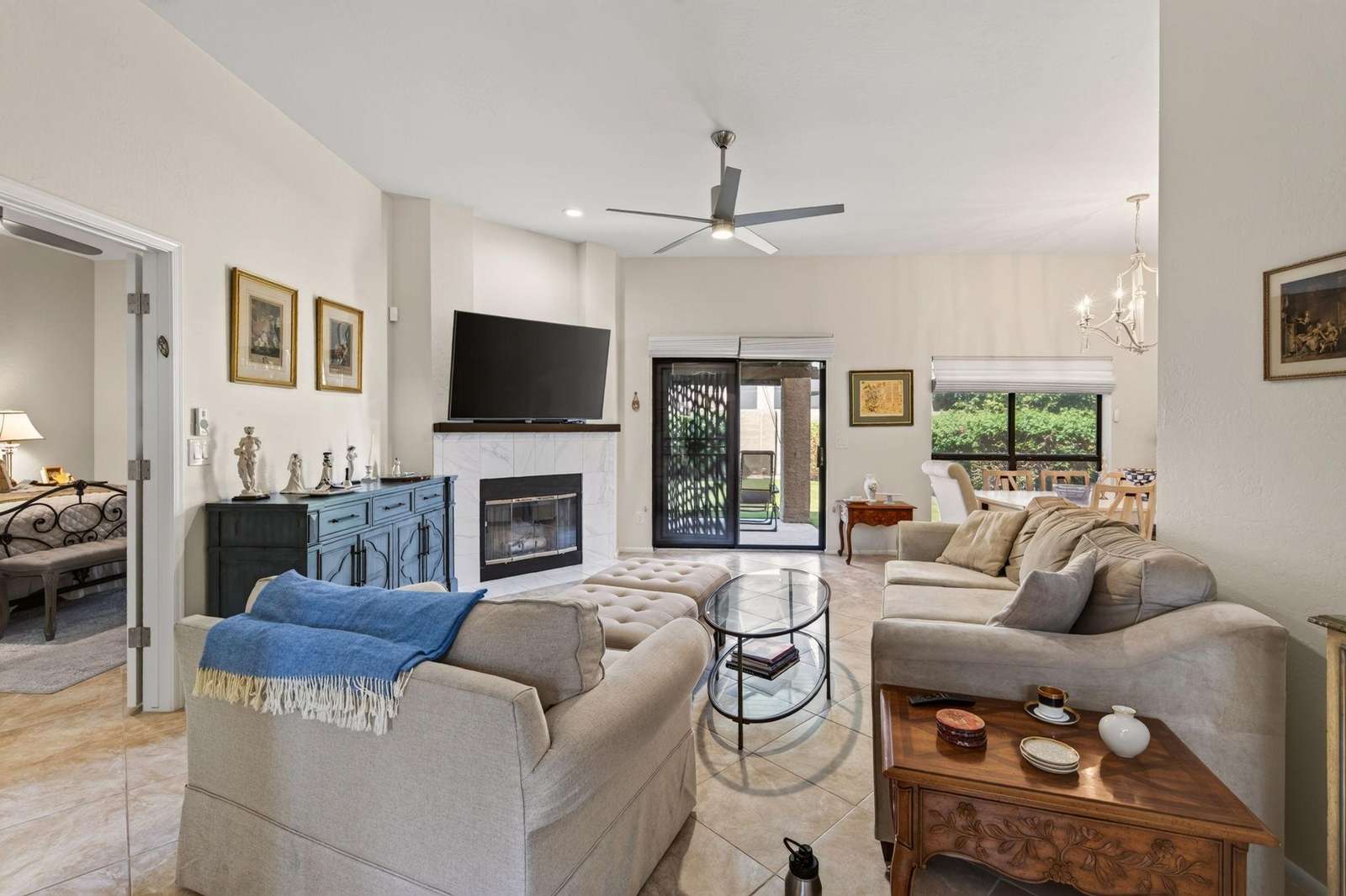 Living room with beige sofas, tufted ottoman, glass coffee table, white marble fireplace, and ceiling fan. - property