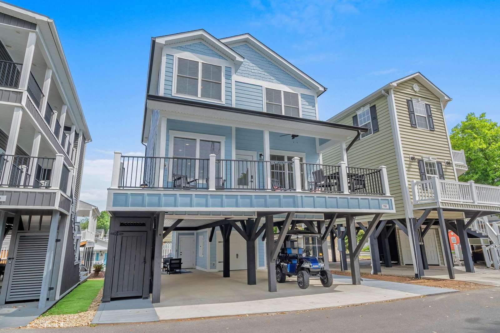 Elevated blue beach house with wraparound balcony, black railings, and covered parking beneath including a golf cart. - property