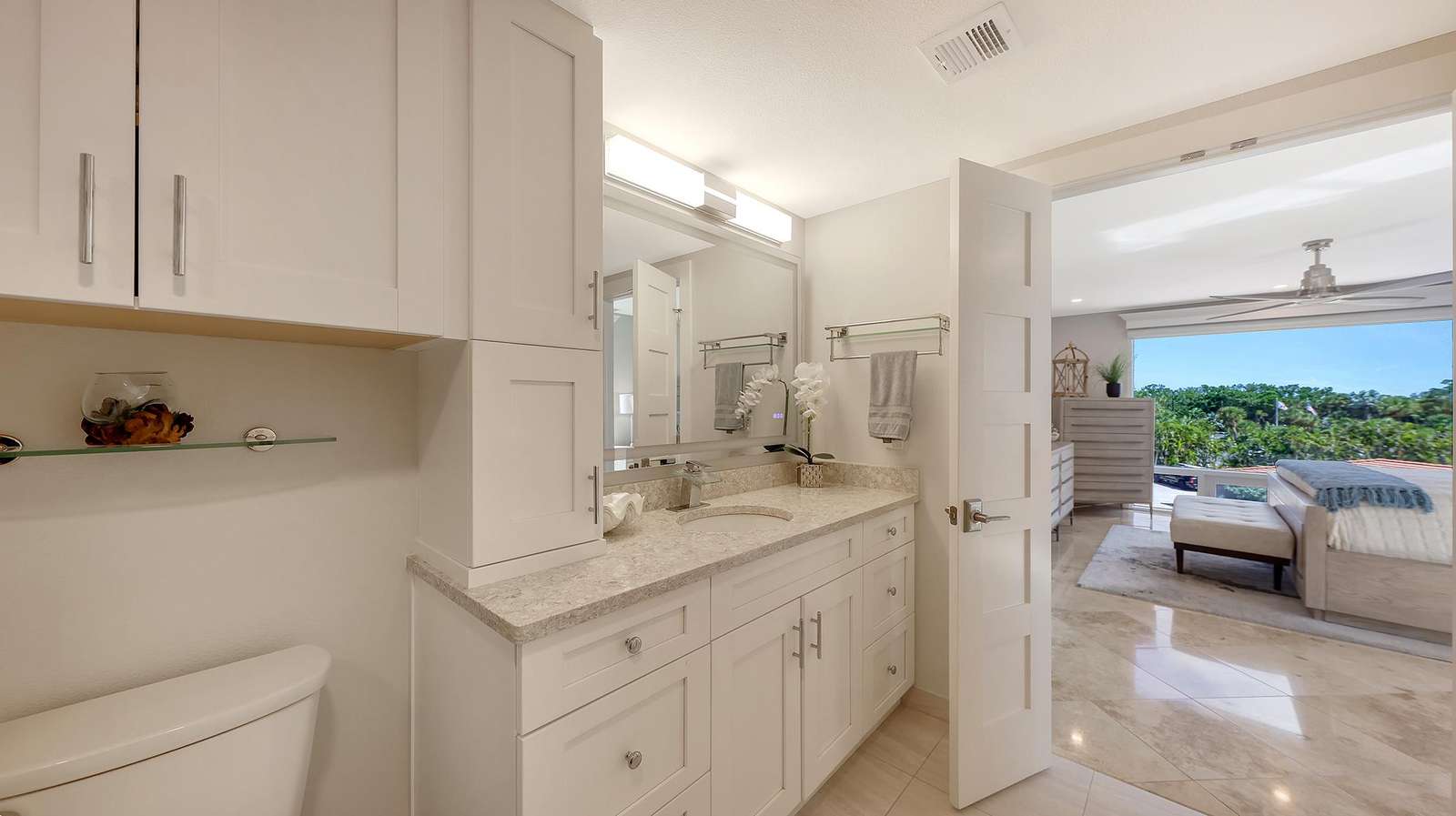 In Suite bathroom vanity with granite countertop, white cabinetry, and a glass shelf above the toilet, open to bedroom.