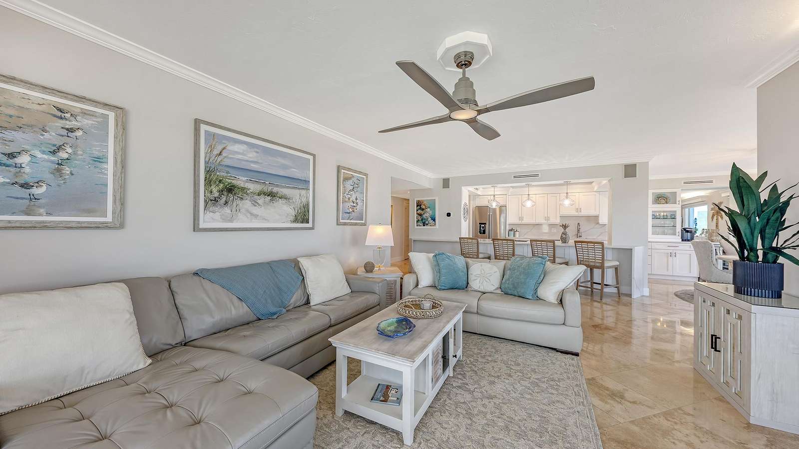 Living room with taupe leather sectional, coastal artwork, whitewashed coffee table, and marble tile flooring.