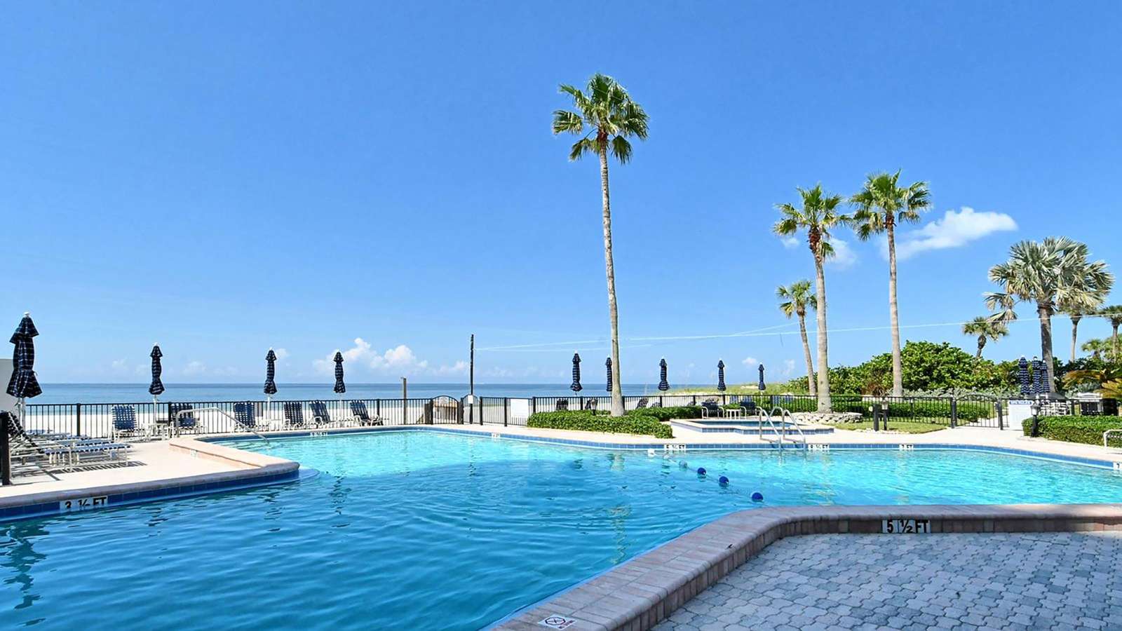 Outdoor pool with blue-tiled edge, surrounded by lounge chairs and closed navy umbrellas, ocean visible beyond.