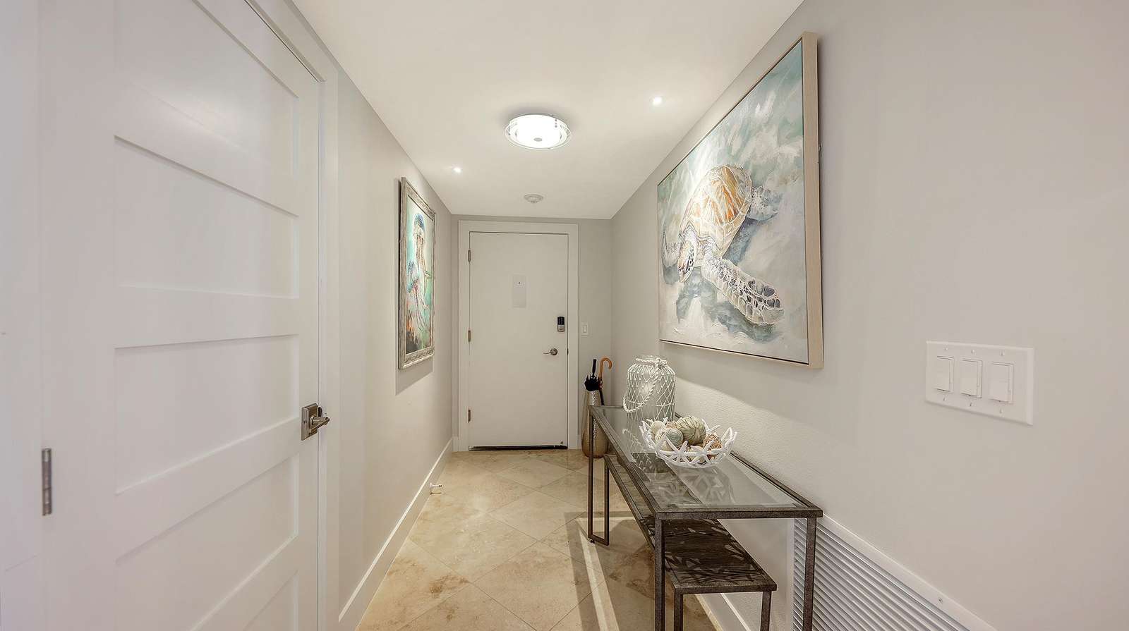 Narrow entry hallway with beige tile floor, glass-top console table, and sea turtle artwork on walls.