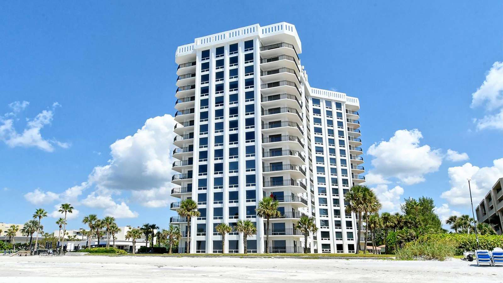 White high-rise condo with wraparound balconies overlooks sandy beach and palm trees under blue sky.