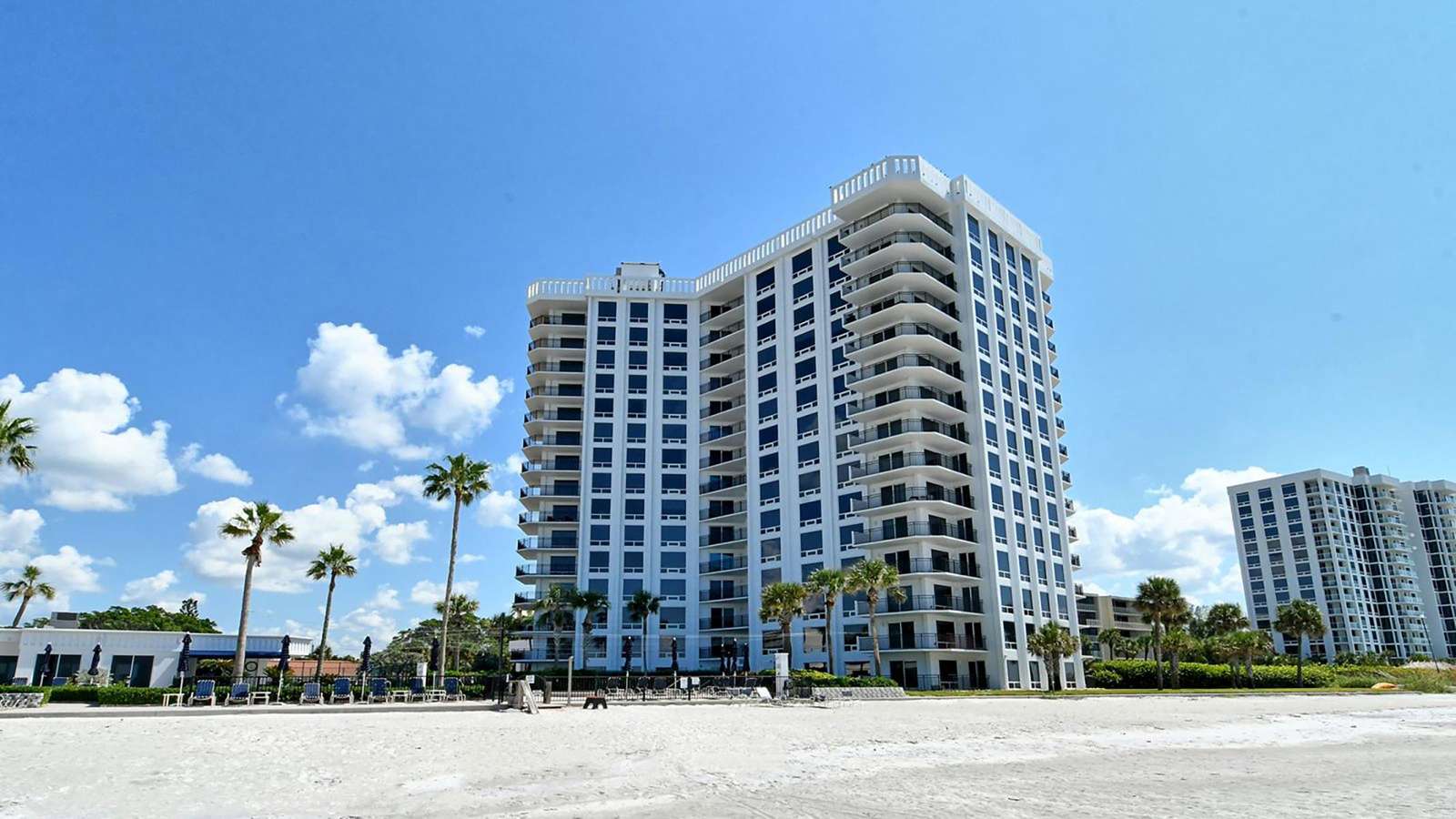 White multi-story condominium building with wraparound balconies against a blue sky, viewed from the sandy beach.