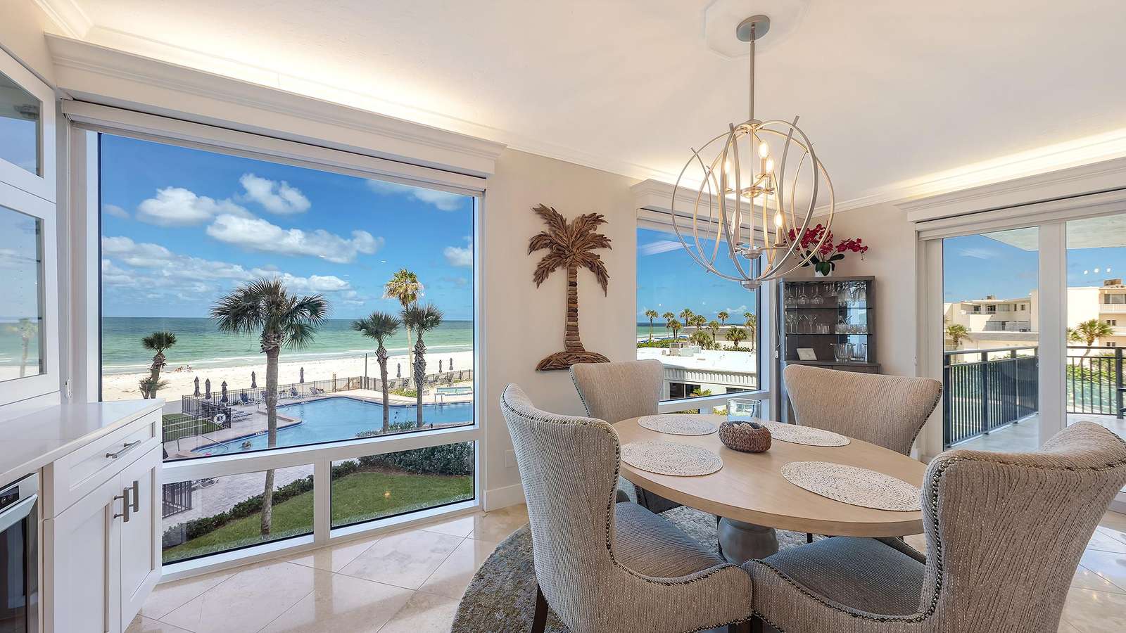 Dining area with round wooden table and four textured chairs, overlooking pool, beach, and ocean through large windows.