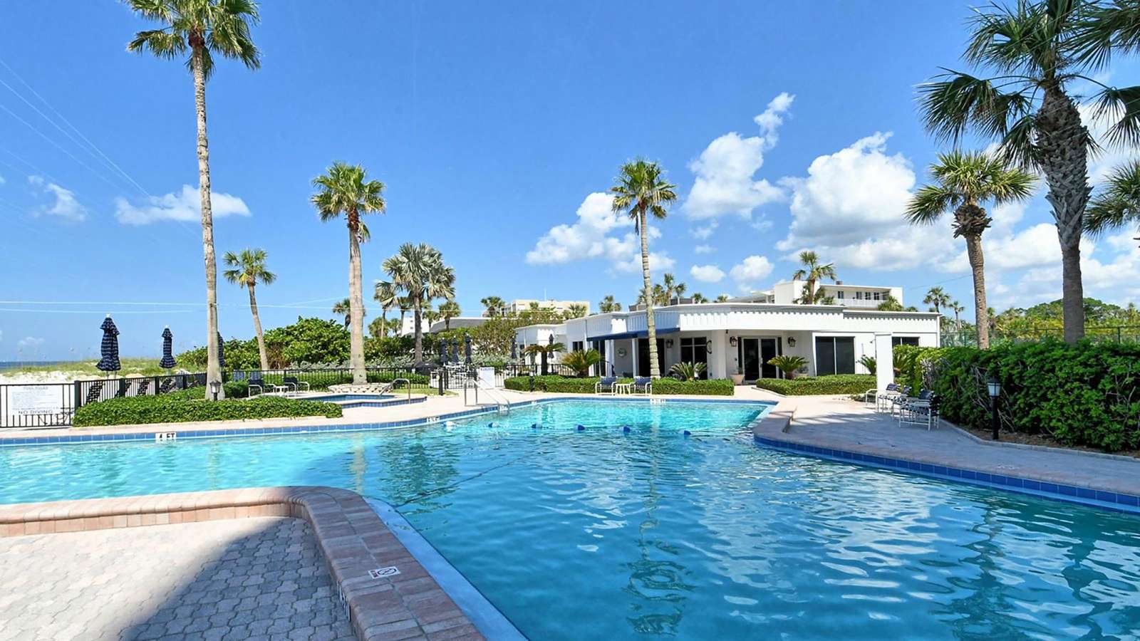 Outdoor pool area with clear blue water, brick-paved deck, palm trees, and a white clubhouse under a bright sky.