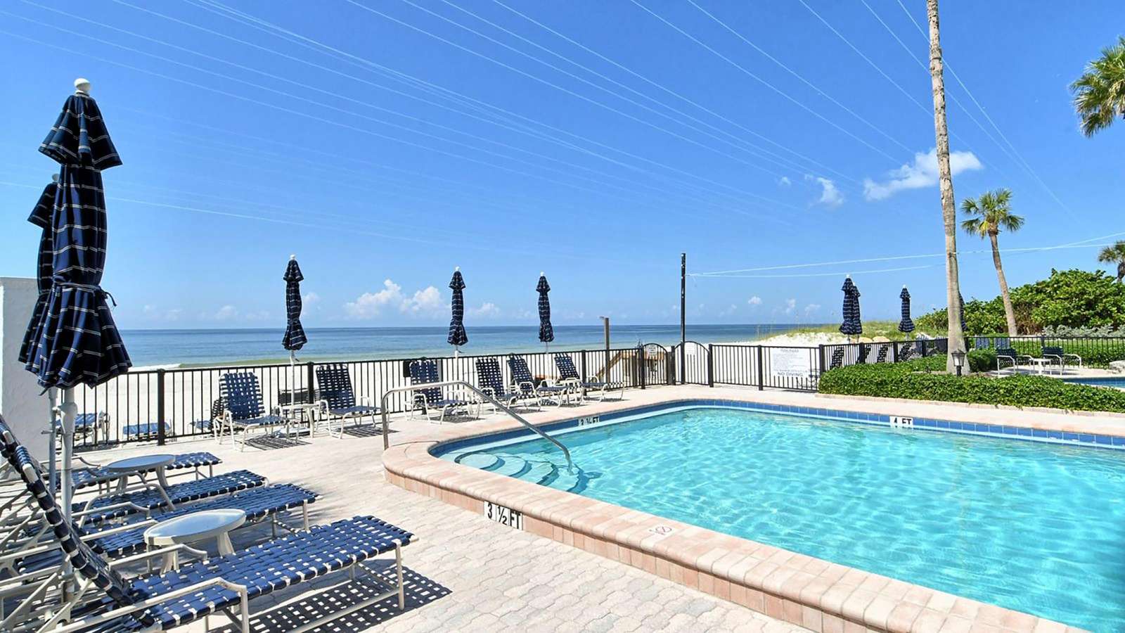 Blue woven lounge chairs and umbrellas surround the rectangular pool with oceanfront views beyond the black metal fence.