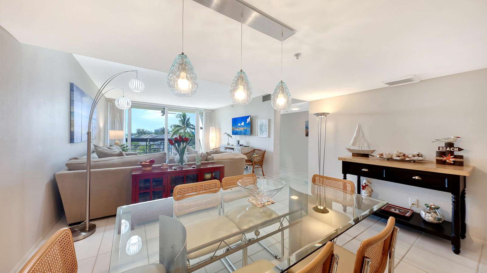 Glass dining table with rattan-back chairs beneath three textured pendant lights and coastal-themed console table.