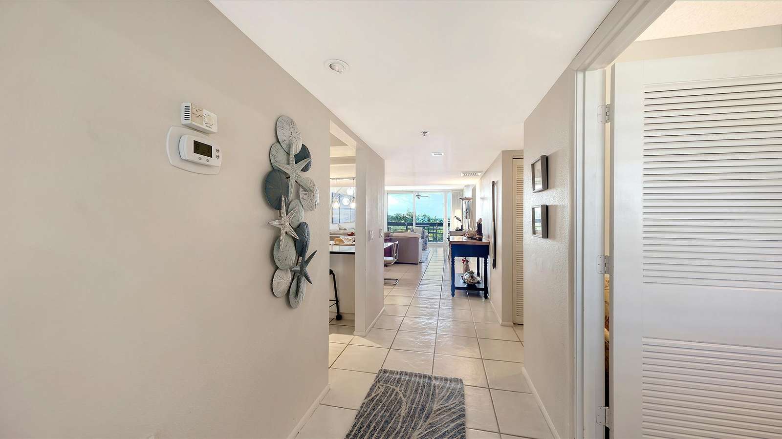 Hallway with beige walls, decorative starfish wall art, white tile flooring, and a gray patterned runner rug.