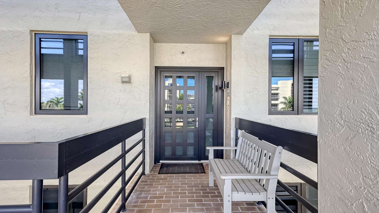 Third-floor entryway with textured cream stucco walls, black metal railing, and a gray wooden bench.