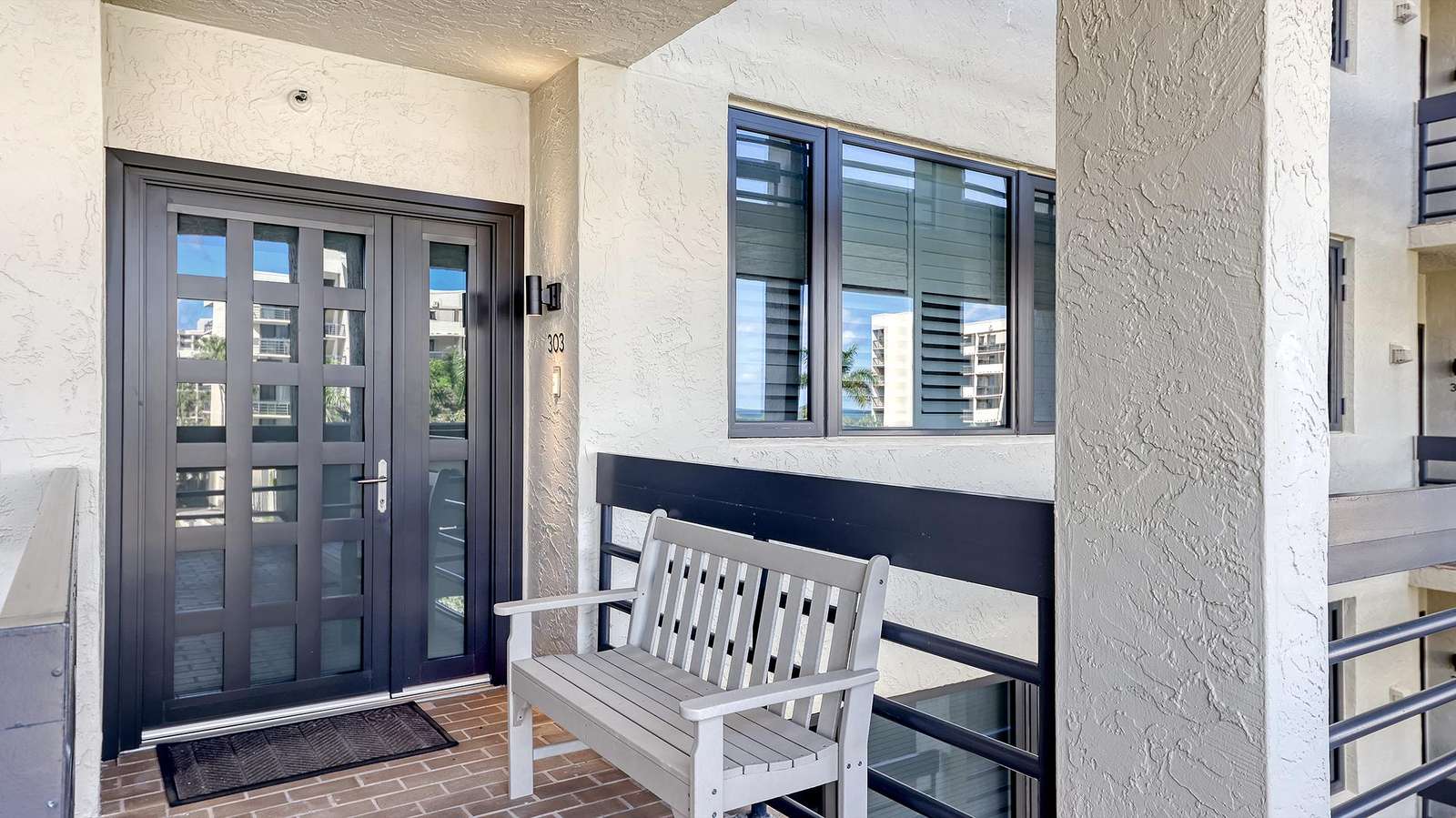 Entryway features double dark wood doors, textured cream walls, and a gray bench on brick flooring.