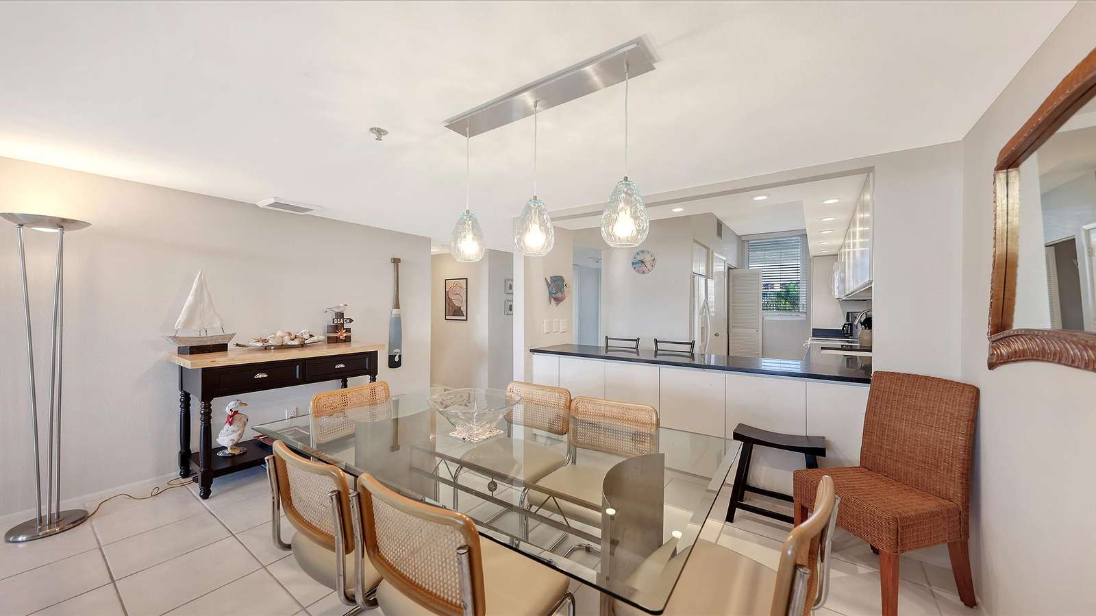 Glass-top dining table with six cane-back chairs under three clear pendant lights, adjacent to a black console table.