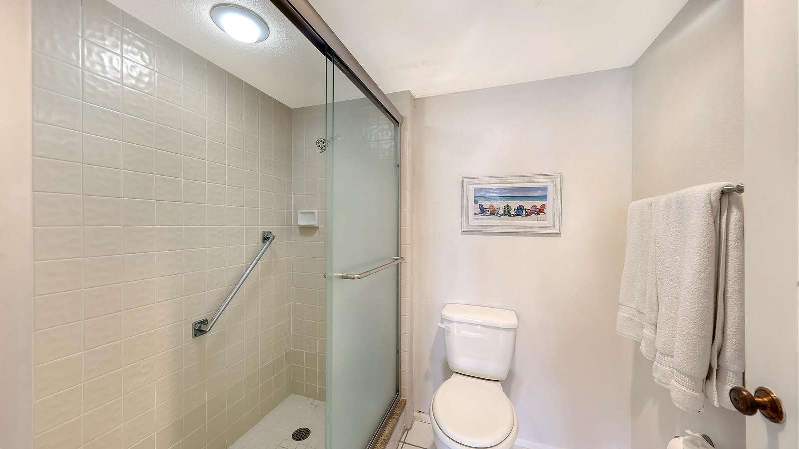 Bathroom features tiled walk-in shower with sliding glass door and white toilet beneath beach-themed wall art.