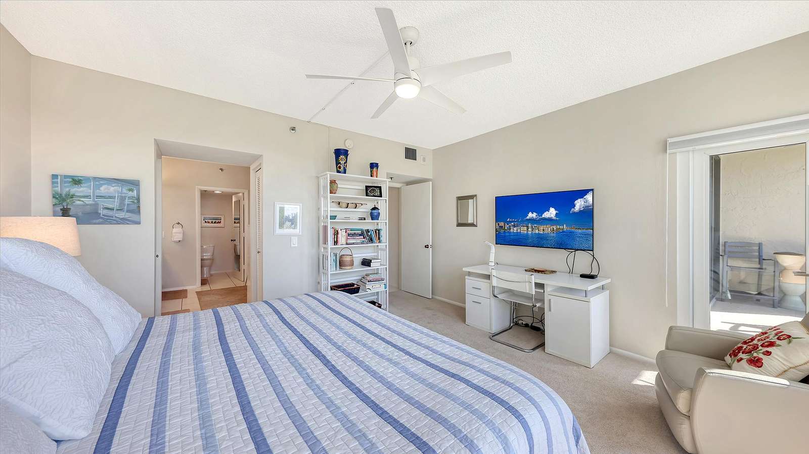 Bedroom with striped blue quilt, white desk under mounted TV, beige armchair, and balcony access.