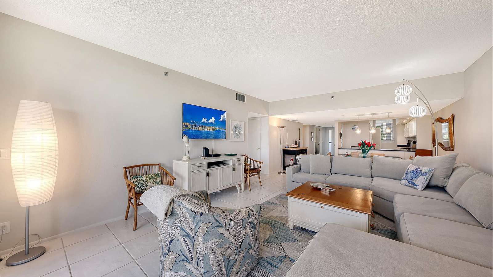 Living area with a large light gray sectional, patterned armchair, white tile flooring, and wall-mounted TV.