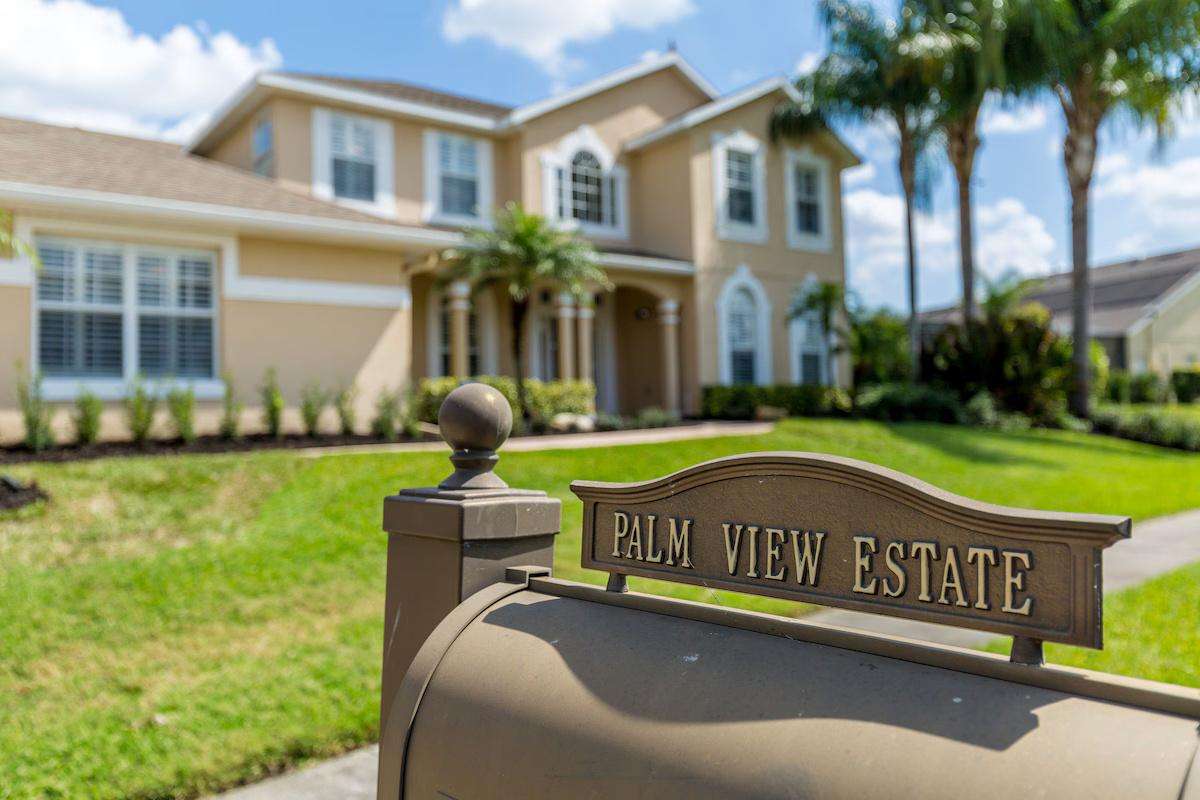 Metal mailbox with property name in front of a two-story beige stucco home and green lawn. - property