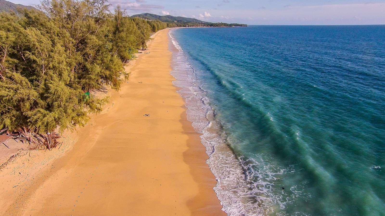 Aerial view of a long sandy beach bordered by green trees and turquoise ocean waves under a clear sky. - property