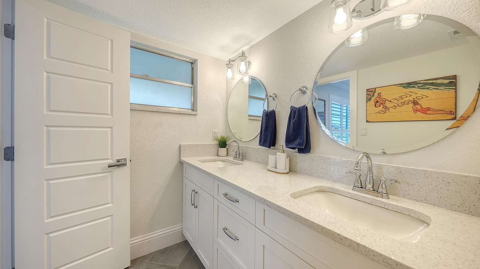 Bathroom features a white quartz double vanity, oval mirrors, chrome fixtures, and a frosted window.
