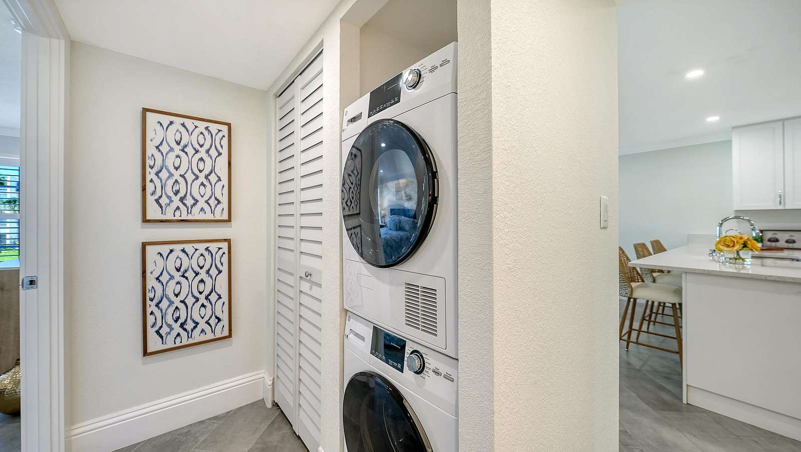 Stacked washer and dryer set in a compact laundry nook with white louvered doors and gray tile flooring.