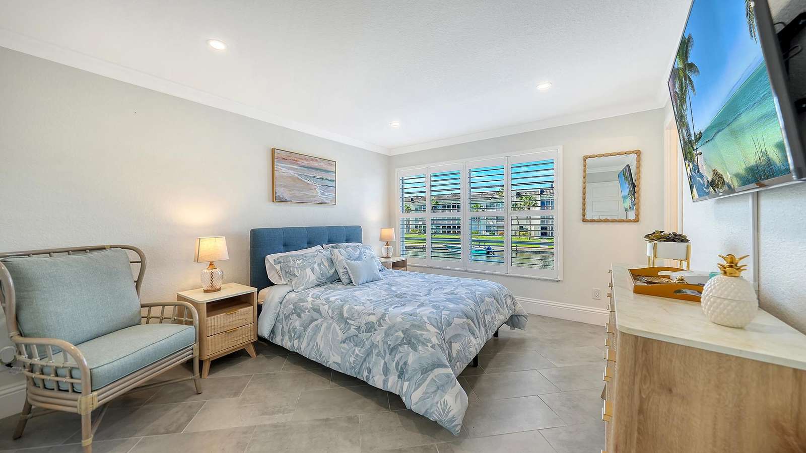 Bedroom with blue upholstered bed, tropical print bedding, wicker nightstand, and water views through plantation shutters.