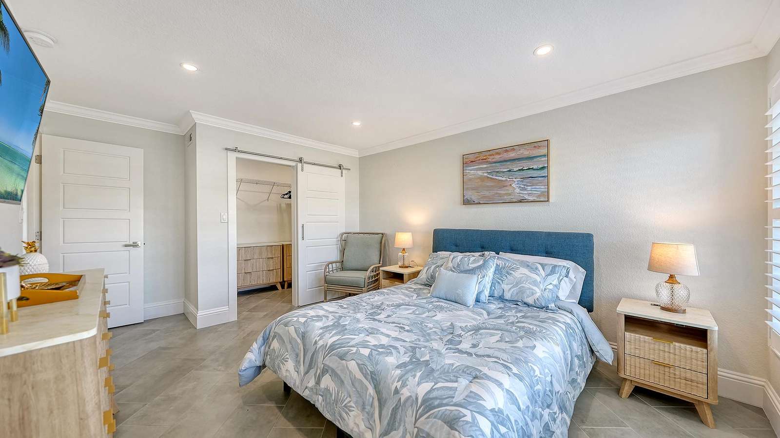 Bedroom with blue upholstered headboard, tropical-patterned bedding, sliding barn door closet, and coastal artwork.