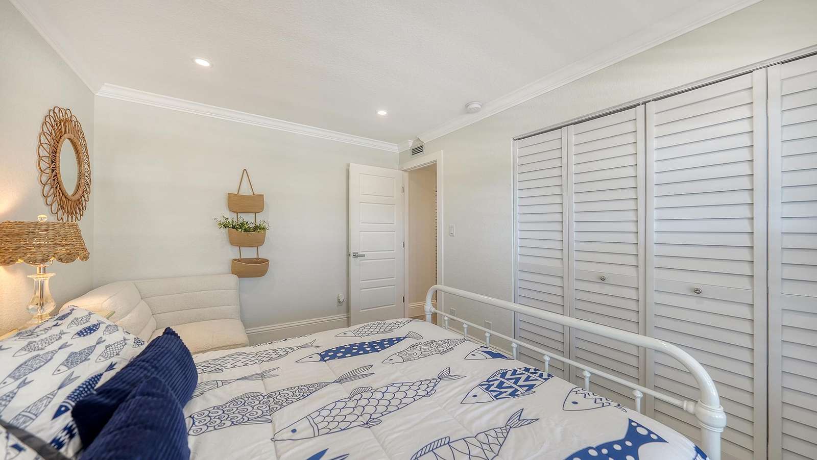 Second Bedroom featuring a white metal bed frame, fish-patterned bedding, white louvered closet doors, and a woven wall mirror.