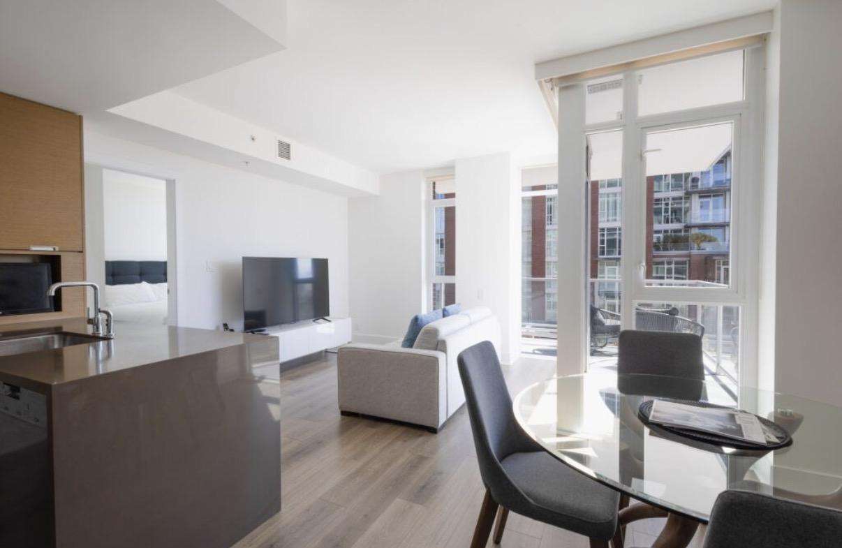 Sunlit living area with gray sofa, wall-mounted TV, glass dining table, and balcony doors revealing city views. - property