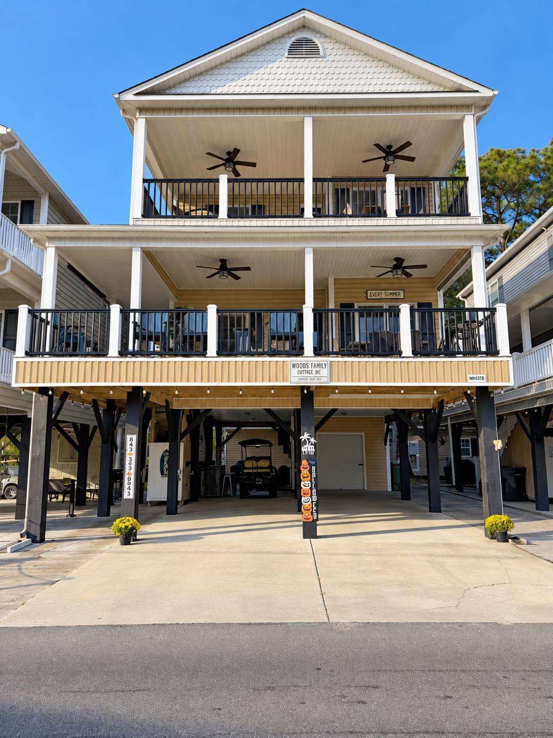 The cottage exterior features two covered balconies with black railings, ceiling fans, white trim, and a shaded ground-level parking area. - property