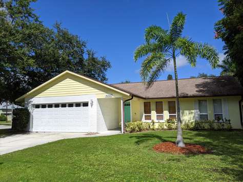 Single-story house with pale yellow siding, white garage door, green front door, and a palm tree on the lawn.