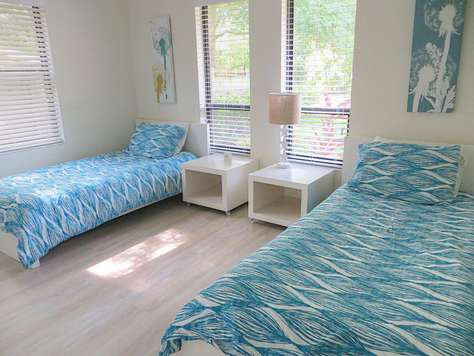 Twin beds with blue wave-patterned bedding flank white nightstands topped with a beige lamp by large windows.