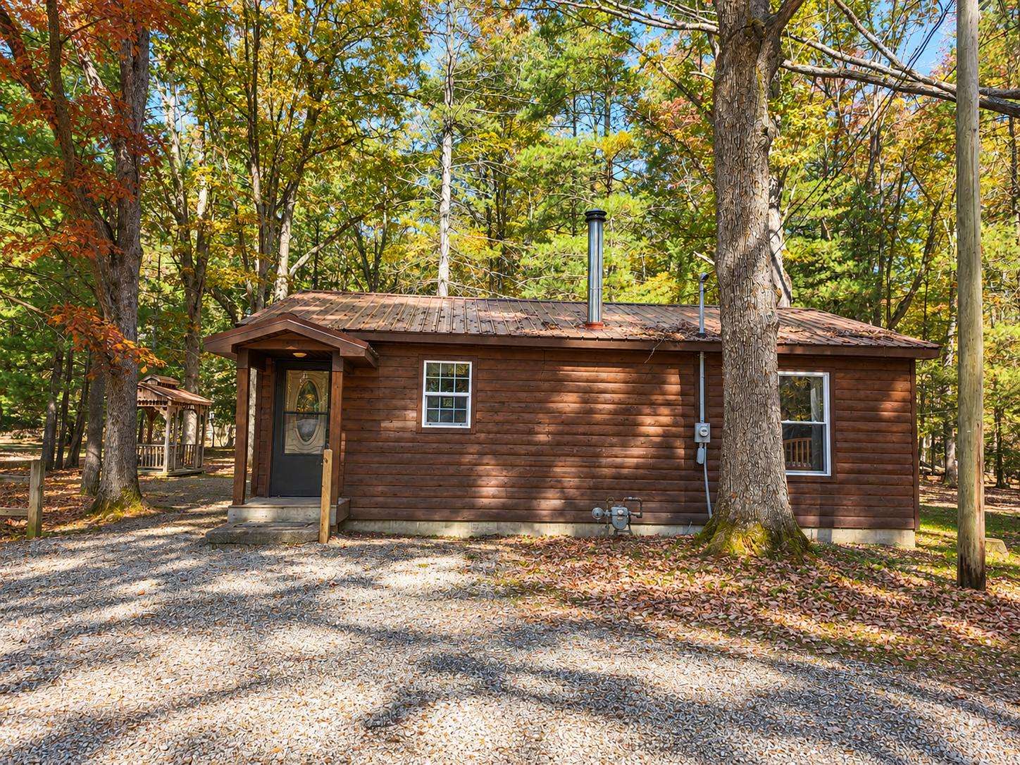 Exterior view of the cabin with dark wood siding, metal roof, and gravel driveway surrounded by autumn trees. - property