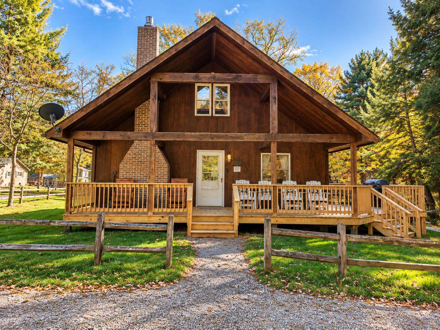 Front view of the cabin features a wide wooden porch with Adirondack chairs, a brick chimney, and rustic fence. - property