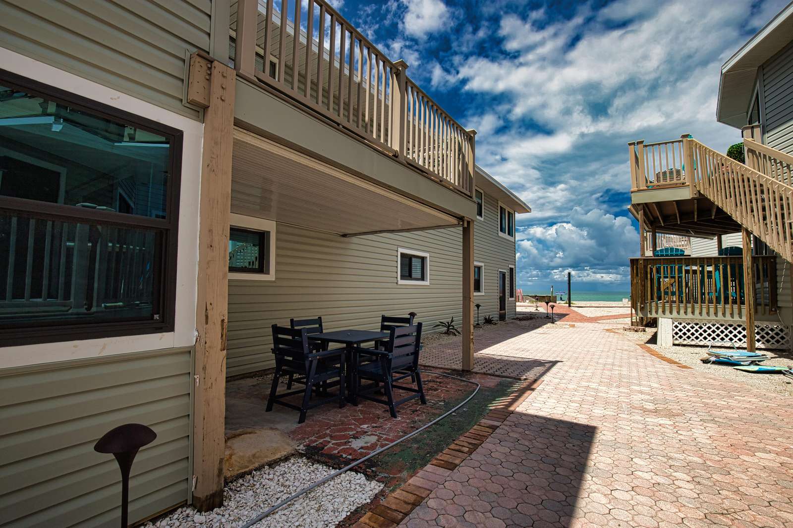 Shaded patio with black wooden table and chairs overlooks a brick-paved walkway leading to the ocean.