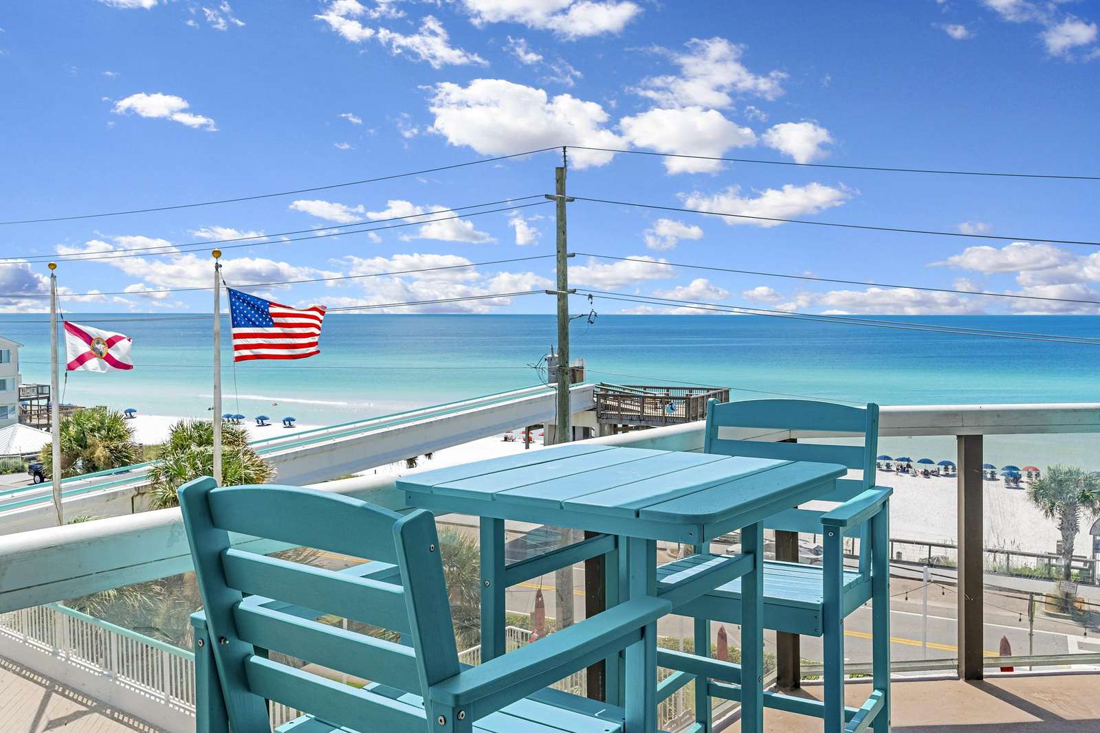 Balcony with turquoise wooden table and chairs overlooks the white sandy beach and blue ocean under a bright sky. - property