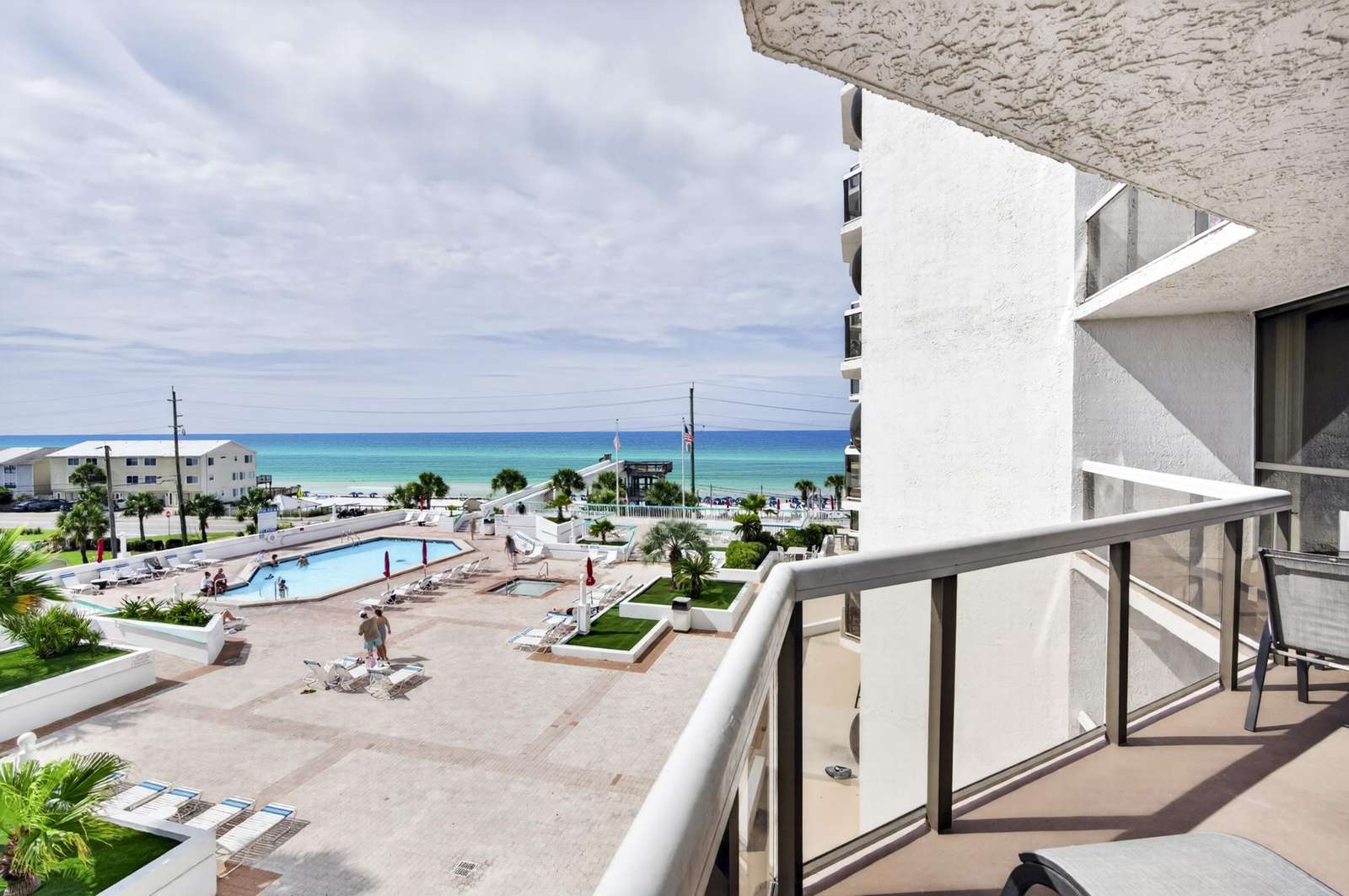 Glass-paneled balcony railing overlooks pool deck, palm trees, and turquoise ocean under a cloudy sky. - property