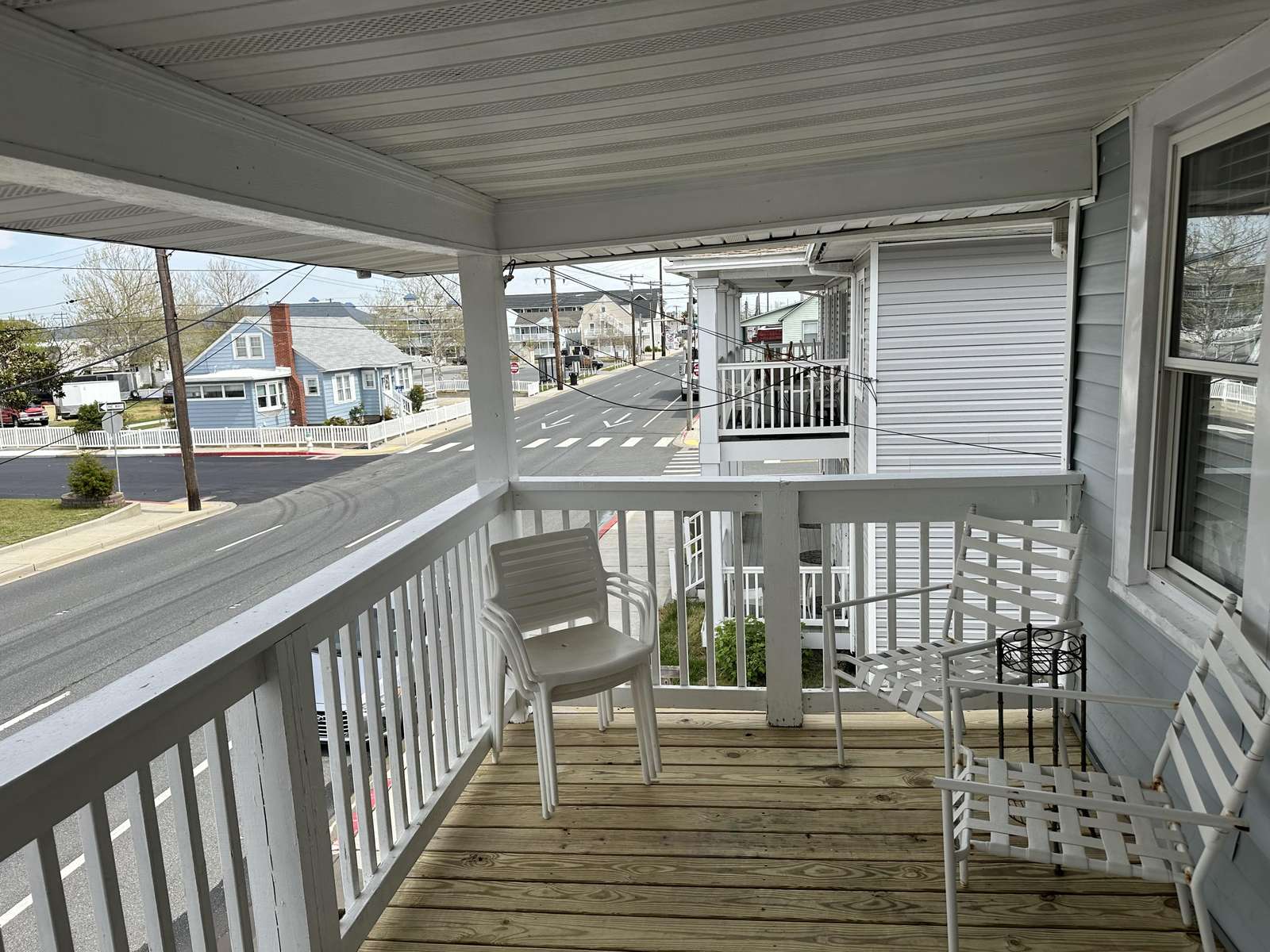 Second-floor covered porch with natural wood decking, white railings, and white metal and plastic chairs overlooking a quiet street.