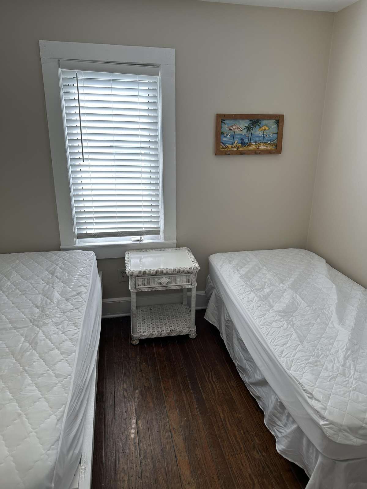 Two twin beds with white quilted covers flank a wicker nightstand beneath a window with white blinds.
