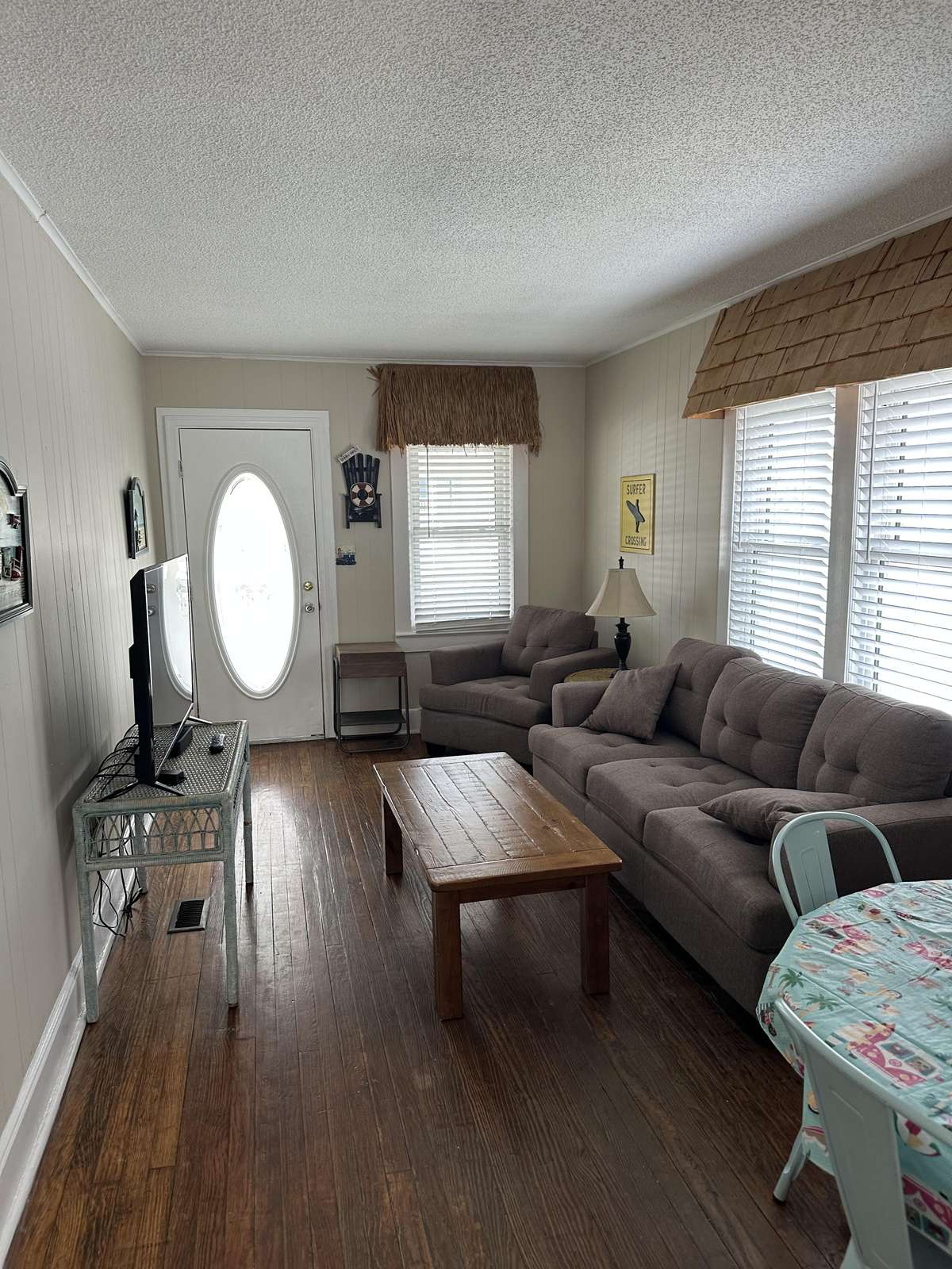 Living room features dark wood floors, gray tufted sofa and armchair, wooden coffee table, and large windows with blinds.
