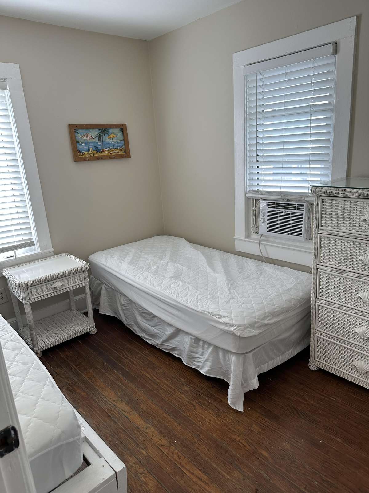Twin beds with white wicker nightstand and dresser sit on dark hardwood floors beneath two windows with blinds.