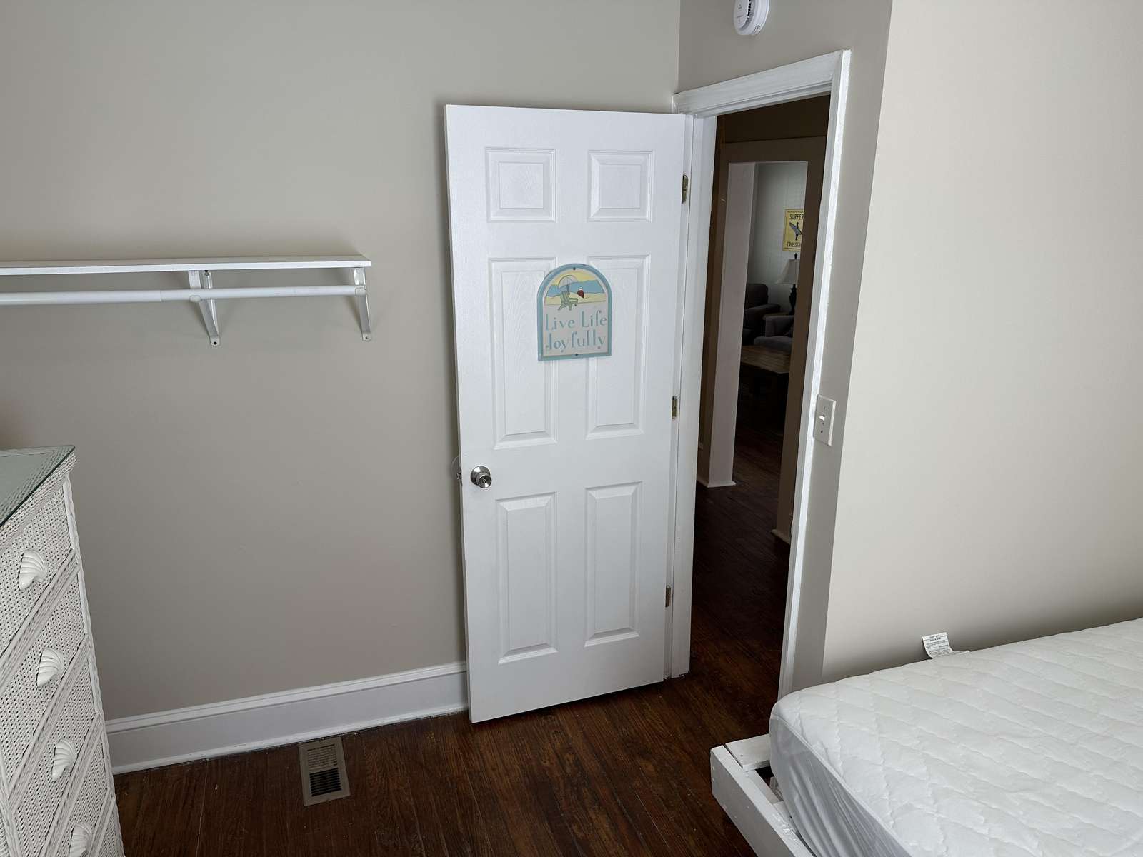 Bedroom corner with white wicker dresser, open six-panel door, hardwood floors, and light gray walls.